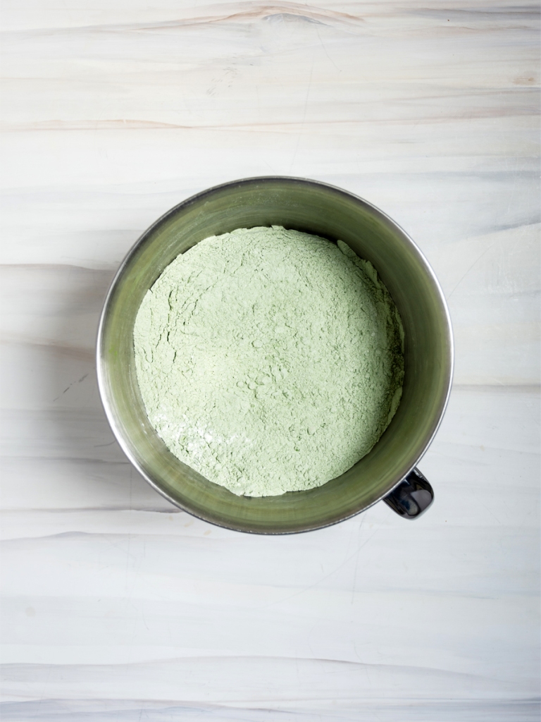 dry ingredients for matcha mochi cake: glutinous rice flour, matcha, sugar, and salt, stirred together in a stainless steel stand mixer bowl on a marble-patterned background
