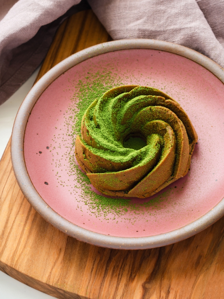 one matcha mochi mini bundt cake on a pink ceramic plate, bundt cake is dusted with matcha with pale lavender linen napkin, cutting board on a pale pink background