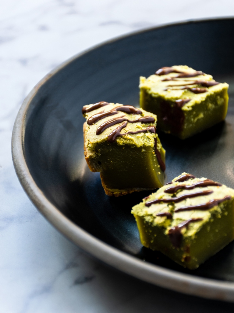 three matcha mochi cubes with drizzled with dark chocolate on a dark ceramic plate and a marble-patterned surface