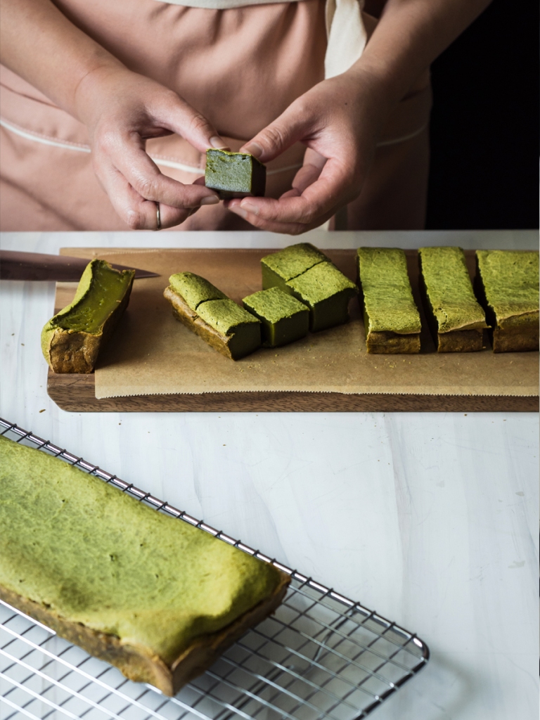 hands holding a cut matcha mochi cube, with various matcha mochi pieces and loaf in frame