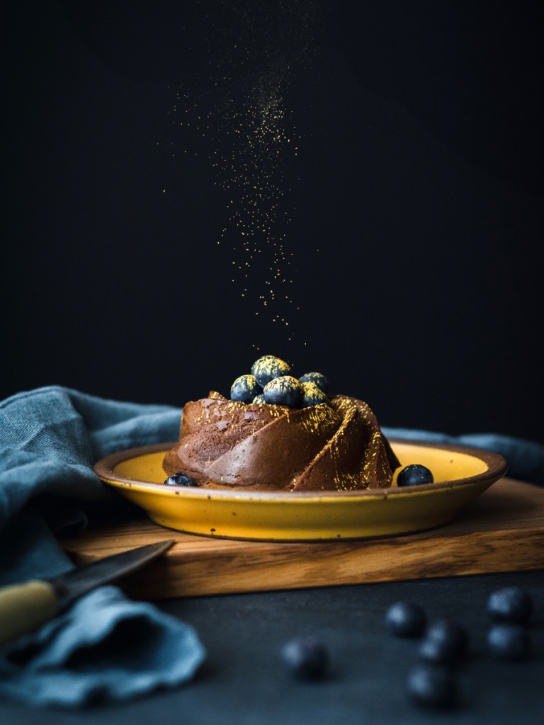 one hojicha mochi mini bundt cake on a yellow ceramic plate, bundt cake is topped with edible gold dust and blueberries against a dark background, with blue linen napkin, cutting board and more blueberries in the foreground