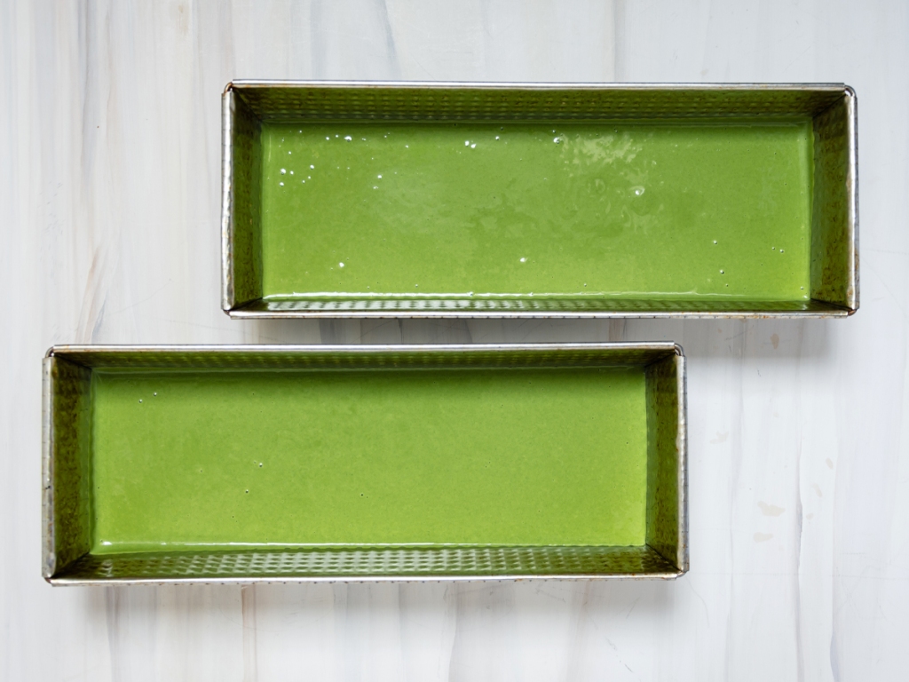 matcha mochi cake batter, divided between two loaf pans on a marble-patterned background